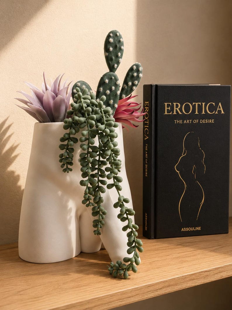 White Uncircumcised Monster Dong planter with succulents, cactus and string of pearls beside a book